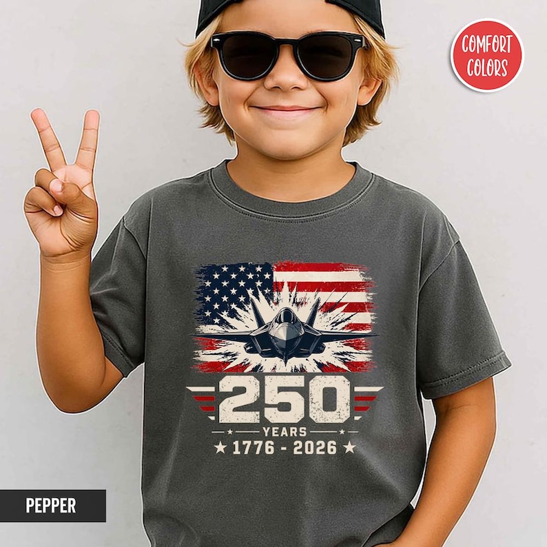 May include: A gray t-shirt featuring a graphic of an American flag and a fighter jet. The design includes the text "250 YEARS 1776-2026". The shirt is worn by a person wearing sunglasses and a black cap.