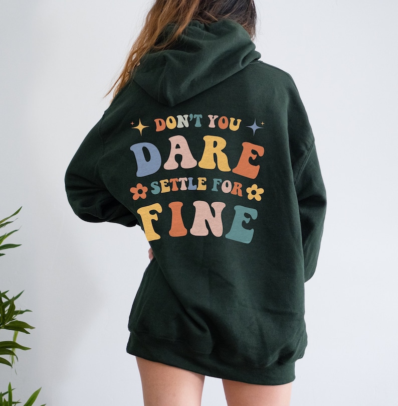Puede incluir: Una sudadera con capucha verde oscuro con el texto "Don't you dare settle for fine" en letras de colores en la espalda.