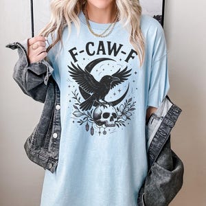 May include: Light blue t-shirt with the text "F-CAW-F" above a black raven graphic perched on a skull, set against a crescent moon and floral elements. The shirt has a relaxed fit.