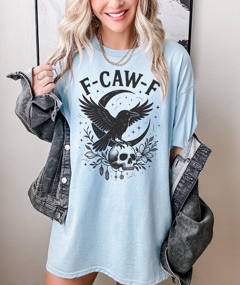 May include: Light blue t-shirt with the text "F-CAW-F" above a black raven graphic perched on a skull, set against a crescent moon and floral elements. The shirt has a relaxed fit.