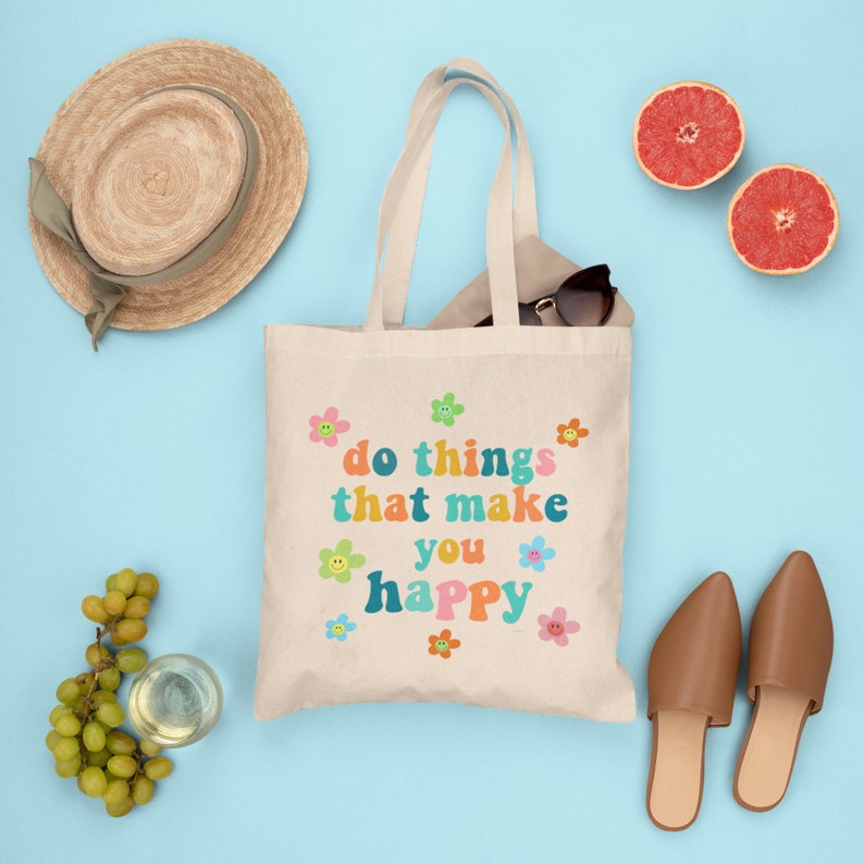 May include: A beige canvas tote bag with the text "do things that make you happy" in colorful retro font. The bag is surrounded by various items, including a straw hat, sunglasses, grape clusters, a glass of white wine, and two pairs of brown leather shoes.