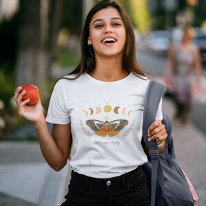 K&ouml;nnte beinhalten: Wei&szlig;es T-Shirt mit einem Schmetterlingsmotiv und dem Text "seek magic everyday". Der Schmetterling hat orange, braune und schwarze Fl&uuml;gel und ist von einem Halbmond und Sternen umgeben.