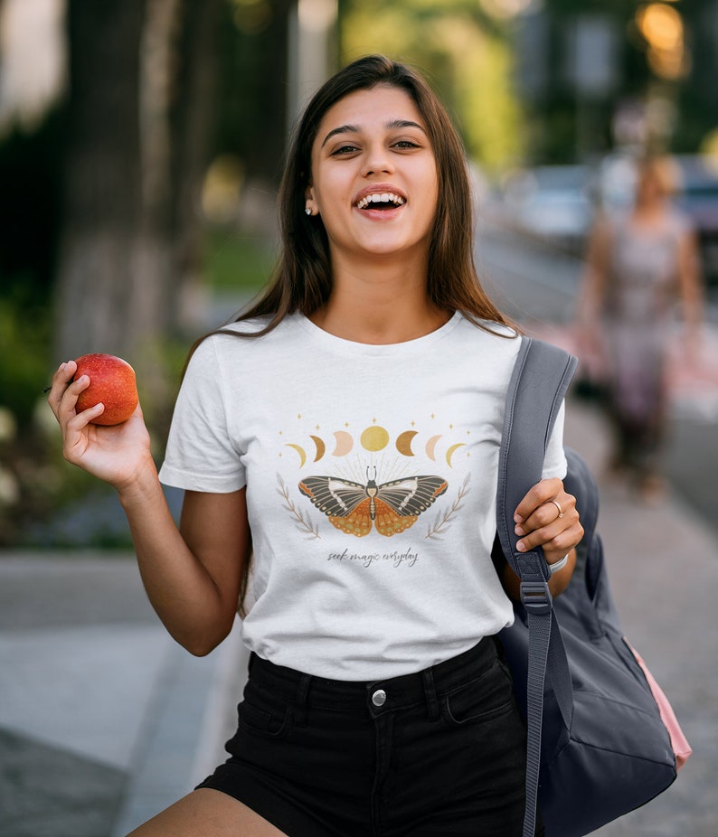 K&ouml;nnte beinhalten: Wei&szlig;es T-Shirt mit einem Schmetterlingsmotiv und dem Text "seek magic everyday". Der Schmetterling hat orange, braune und schwarze Fl&uuml;gel und ist von einem Halbmond und Sternen umgeben.
