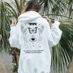 May include: White hoodie with a black graphic design featuring a butterfly, hands, an eye, and the text "444 The universe and your spiritual guides are protecting you."