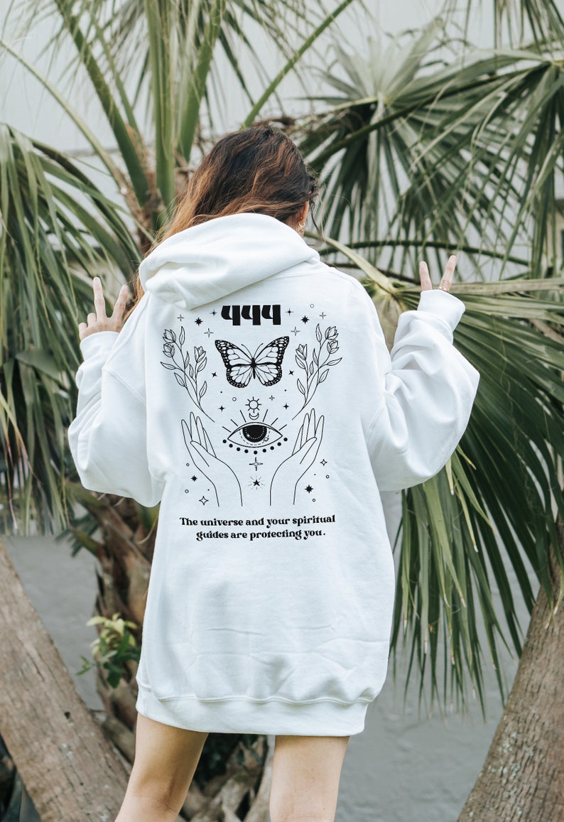 May include: White hoodie with a black graphic design featuring a butterfly, hands, an eye, and the text "444 The universe and your spiritual guides are protecting you."