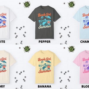 K&ouml;nnte beinhalten: Sechs T-Shirts in verschiedenen Farben mit einem Motiv einer Frau, die am Strand ein Buch liest, mit dem Text "Book Girl Summer". Die Farben sind Wei&szlig;, Pfeffer, Chambray, Elfenbein, Banane und Bl&uuml;te.