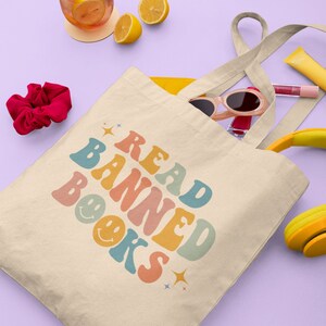 Peut inclure: Un sac fourre-tout beige avec le texte "READ BANNED BOOKS" en police r&eacute;tro color&eacute;e. Le texte est entour&eacute; d'&eacute;toiles et de visages souriants. Le sac est rempli de divers articles, notamment des lunettes de soleil, un &eacute;lastique &agrave; cheveux et un casque jaune et noir.