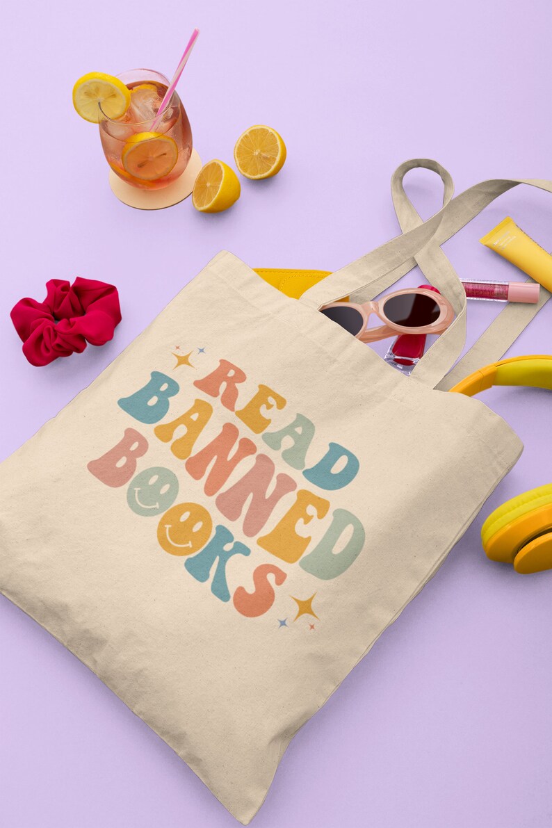 Peut inclure: Un sac fourre-tout beige avec le texte "READ BANNED BOOKS" en police r&eacute;tro color&eacute;e. Le texte est entour&eacute; d'&eacute;toiles et de visages souriants. Le sac est rempli de divers articles, notamment des lunettes de soleil, un &eacute;lastique &agrave; cheveux et un casque jaune et noir.