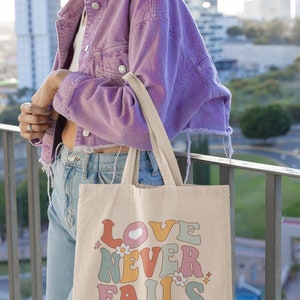 May include: A beige tote bag with a colorful retro design that says "LOVE NEVER FAILS" with a heart and flowers. The text "1 Corinthians 13:8" is printed below the design.