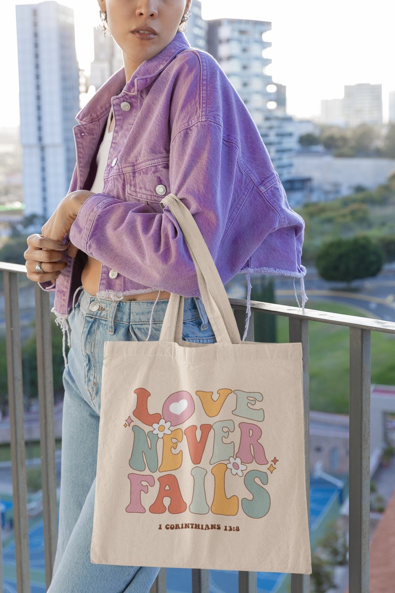 May include: A beige tote bag with a colorful retro design that says "LOVE NEVER FAILS" with a heart and flowers. The text "1 Corinthians 13:8" is printed below the design.