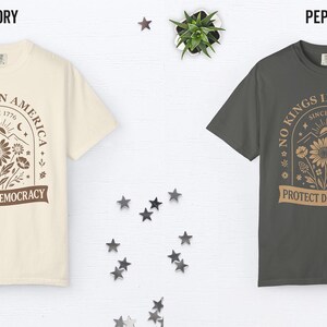 May include: Two t-shirts, ivory and pepper, are displayed. Each shirt features a graphic with the text "NO KINGS IN AMERICA SINCE 1776 PROTECT DEMOCRACY" surrounding a floral design. The shirts are on a white surface with star decorations.