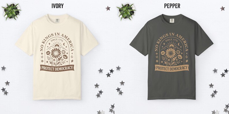 May include: Two t-shirts, ivory and pepper, are displayed. Each shirt features a graphic with the text "NO KINGS IN AMERICA SINCE 1776 PROTECT DEMOCRACY" surrounding a floral design. The shirts are on a white surface with star decorations.