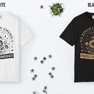 May include: White and black t-shirts with a graphic design. The design includes the text "NO KINGS IN AMERICA SINCE 1776" and "PROTECT DEMOCRACY" with floral and mountain illustrations. The shirts are displayed on a white surface with star decorations.