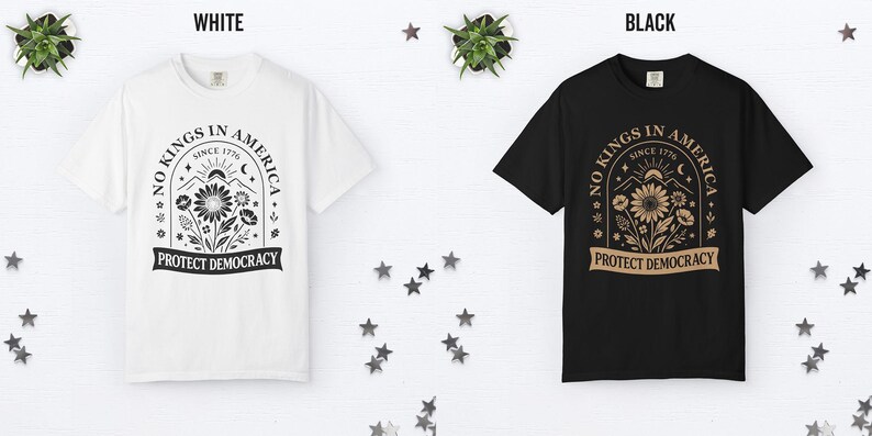 May include: White and black t-shirts with a graphic design. The design includes the text "NO KINGS IN AMERICA SINCE 1776" and "PROTECT DEMOCRACY" with floral and mountain illustrations. The shirts are displayed on a white surface with star decorations.