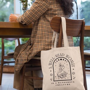 Pode incluir: Saco tote creme com texto preto: "A WELL-READ WOMAN IS A DANGEROUS CREATURE". O design inclui um livro aberto, flores, estrelas e fases da lua. O saco est&aacute; pendurado numa cadeira de madeira.