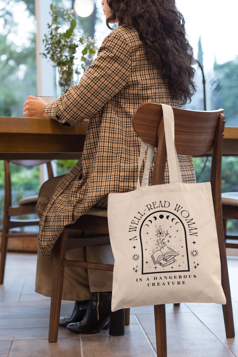 Pode incluir: Saco tote creme com texto preto: "A WELL-READ WOMAN IS A DANGEROUS CREATURE". O design inclui um livro aberto, flores, estrelas e fases da lua. O saco est&aacute; pendurado numa cadeira de madeira.