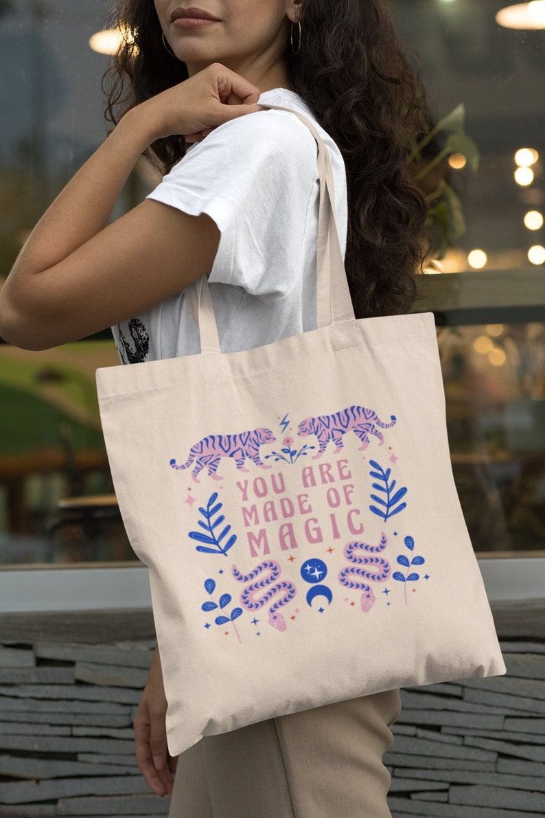 Op de afbeelding: Beige draagtas met een kleurrijk ontwerp. Het ontwerp bevat twee tijgers, de tekst "YOU ARE MADE OF MAGIC", een slang en bloemelementen in roze en blauw. De tas heeft lange beige hengsels.