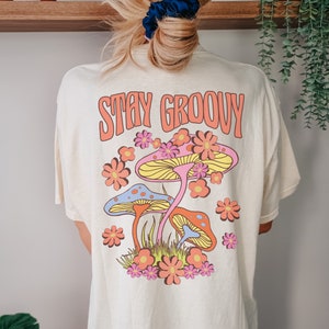 May include: A white t-shirt with a graphic of colorful mushrooms and flowers. The text "STAY GROOVY" is printed in orange on the shirt.
