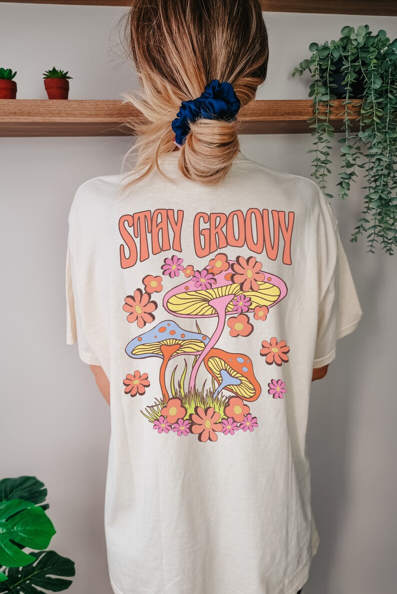 May include: A white t-shirt with a graphic of colorful mushrooms and flowers. The text "STAY GROOVY" is printed in orange on the shirt.