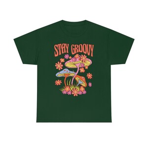 May include: Dark green t-shirt featuring a colorful graphic of mushrooms and flowers. The text "STAY GROOVY" is printed in orange above the illustration. The design includes yellow, pink, orange, and blue elements, creating a vibrant and retro aesthetic.