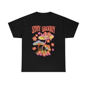 May include: Black t-shirt with a colorful graphic of mushrooms and flowers. The text "STAY GROOVY" is in orange above the illustration. The design features blue, yellow, orange, and pink elements, creating a vibrant and retro aesthetic.