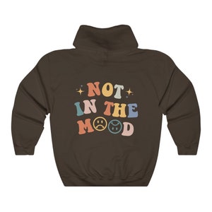 Pu&ograve; includere: Una felpa con cappuccio marrone con il testo "NOT IN THE MOOD" in colori arcobaleno con emoji di facce tristi.