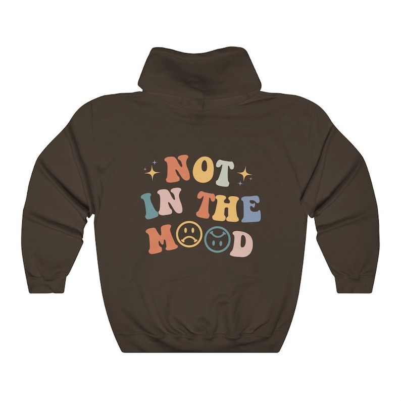 Pu&ograve; includere: Una felpa con cappuccio marrone con il testo "NOT IN THE MOOD" in colori arcobaleno con emoji di facce tristi.
