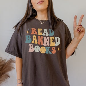 May include: A dark brown t-shirt with the text "READ BANNED BOOKS" in colourful retro letters. The text includes two yellow smiley faces.