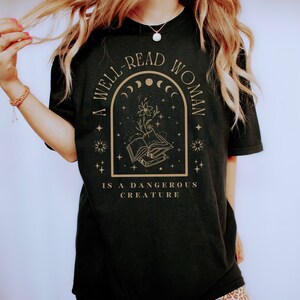 May include: A black t-shirt with a gold graphic design that reads "A well-read woman is a dangerous creature". The graphic features a book, a flower, and stars.