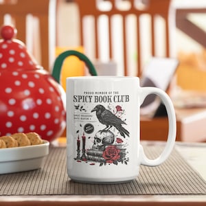 Puede incluir: Taza de cer&aacute;mica blanca con una ilustraci&oacute;n en blanco y negro de un cuervo posado sobre una calavera con una rosa roja. El texto "Proud Member of the Spicy Book Club" est&aacute; impreso en la taza. El texto "Smut Readers' Safe Haven" tambi&eacute;n est&aacute; impreso en la taza.