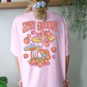May include: Pink t-shirt with the text "Stay Groovy" and a retro graphic of mushrooms and flowers.
