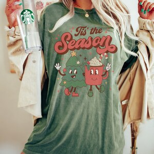 May include: Green t-shirt with a retro-style graphic of a Christmas tree and a mug of hot chocolate. The text "Tis' the Season" is printed in red on the shirt.
