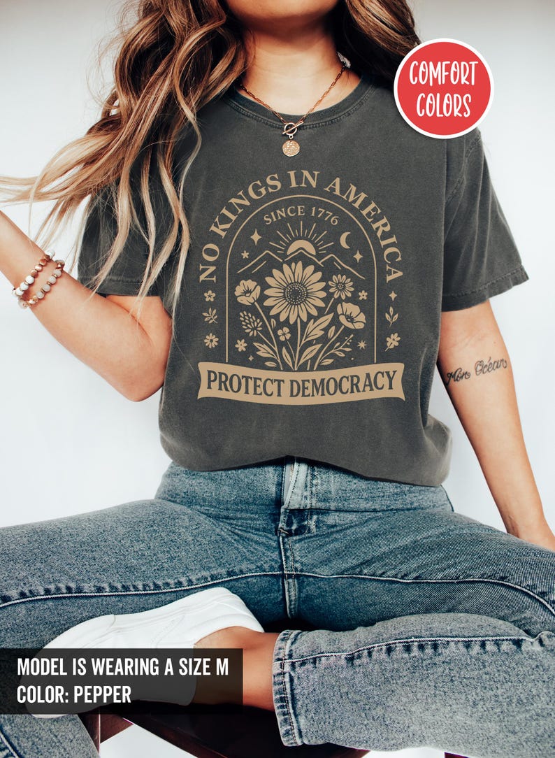 May include: Dark grey t-shirt with the text "NO KINGS IN AMERICA SINCE 1776 PROTECT DEMOCRACY". The shirt features a floral design with a mountain and sun graphic. The shirt is a comfort color.