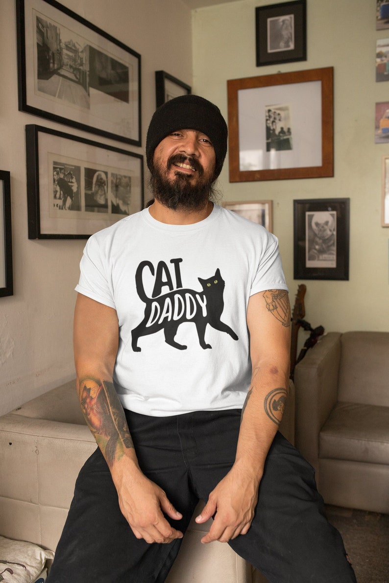 May include: White t-shirt with the text "CAT DADDY" and a black cat silhouette. The cat has yellow eyes. The shirt is worn by a person wearing a black beanie and black pants.