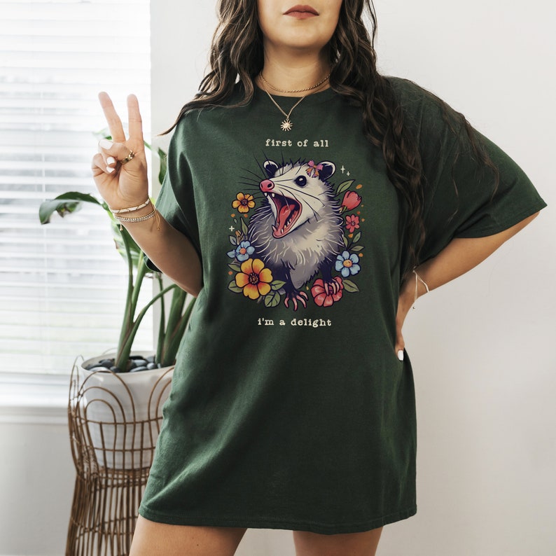 May include: A dark green t-shirt with a graphic of a possum with flowers and the text "first of all I'm a delight".