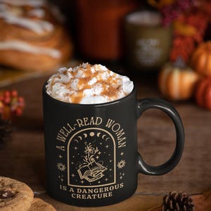 May include: Black ceramic mug filled with a beverage topped with whipped cream and caramel drizzle. The mug features the quote "A Well-Read Woman Is A Dangerous Creature" in gold lettering, with a floral and celestial design.