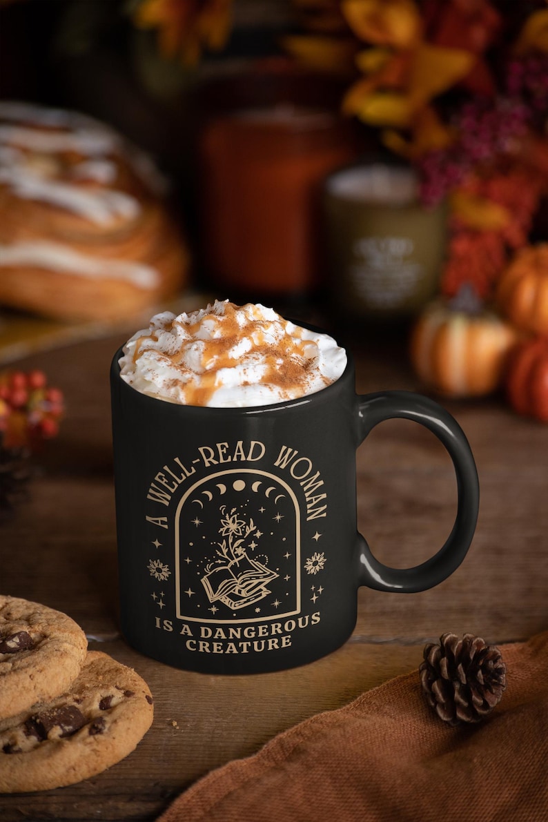 May include: Black ceramic mug filled with a beverage topped with whipped cream and caramel drizzle. The mug features the quote "A Well-Read Woman Is A Dangerous Creature" in gold lettering, with a floral and celestial design.