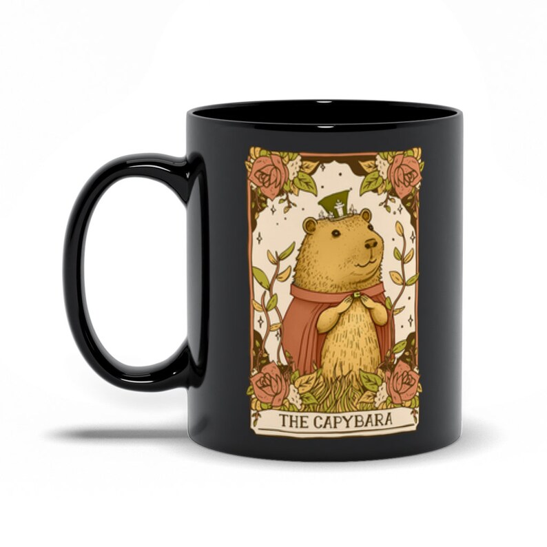 May include: Black coffee mug with a graphic of a capybara wearing a hat and a red cape. The graphic is surrounded by flowers and the text "THE CAPYBARA".