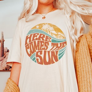May include: A white t-shirt with a retro-style graphic of a sun, waves, and palm trees. The graphic says "Here Comes The Sun".