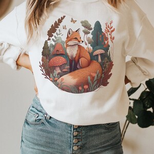 May include: White sweatshirt with a graphic print of a fox in a forest setting. The fox is orange and brown with a bushy tail. The forest is depicted with green trees, mushrooms, and flowers.