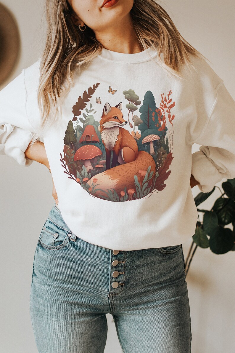 May include: White sweatshirt with a graphic print of a fox in a forest setting. The fox is orange and brown with a bushy tail. The forest is depicted with green trees, mushrooms, and flowers.