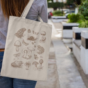 May include: A beige tote bag with a brown illustration of mushrooms, frogs, and butterflies. The illustration is in a vintage style.
