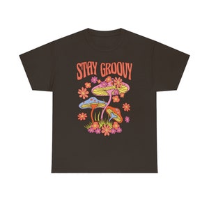 May include: A dark brown t-shirt featuring a colorful graphic of mushrooms and flowers. The text "STAY GROOVY" is printed above the illustration in orange. The design has a retro, psychedelic vibe.