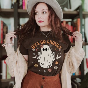 May include: A woman wearing a brown t-shirt with a white ghost graphic and the text "Let's Go Ghouls". She is also wearing a brown skirt, a white cardigan, and a gray fedora.