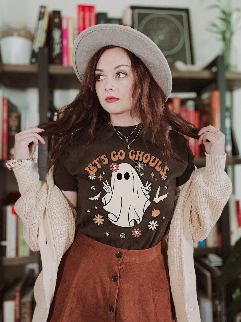 May include: A woman wearing a brown t-shirt with a white ghost graphic and the text "Let's Go Ghouls". She is also wearing a brown skirt, a white cardigan, and a gray fedora.