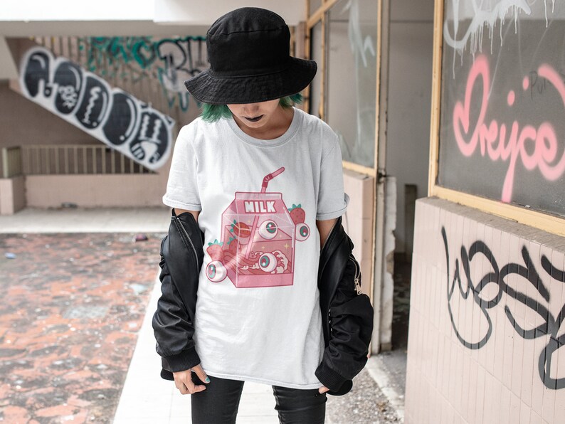 Puede incluir: Una camiseta blanca con una ilustraci&oacute;n de dibujos animados rosa y blanca de una caja de leche con fresas y ojos. El texto "MILK" est&aacute; impreso en la caja.