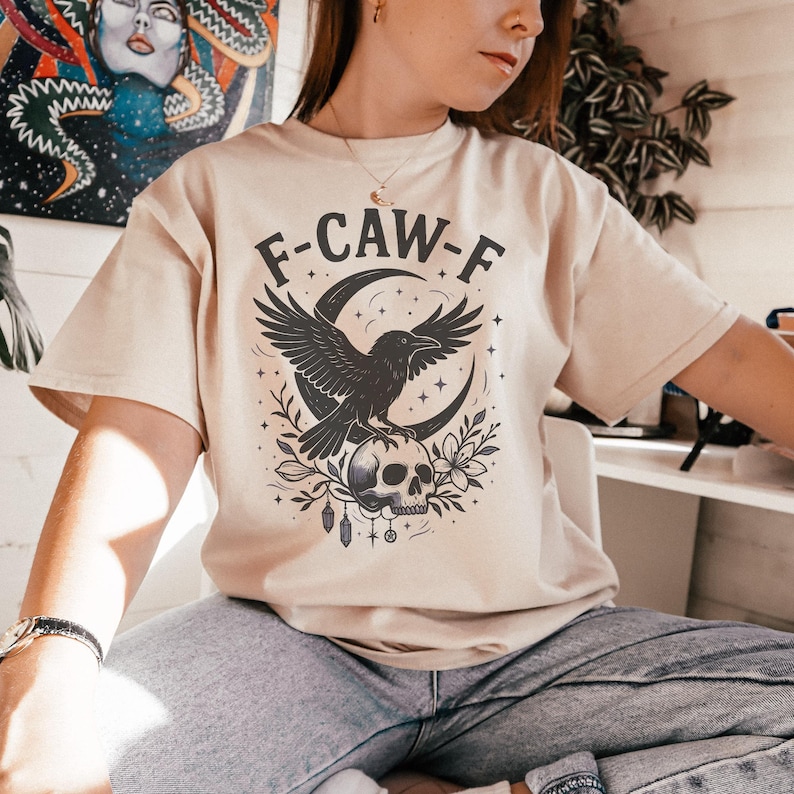 May include: Beige t-shirt with a graphic featuring a black crow perched on a skull, set against a crescent moon. The text "F-CAW-F" is arched above the design. The design includes floral and star elements.