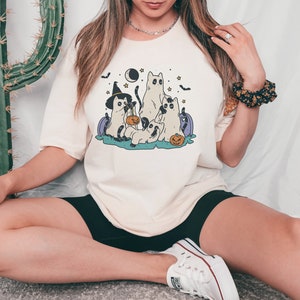May include: A white t-shirt with a graphic of four cats dressed up for Halloween. The cats are wearing witch hats, holding pumpkins, and have stars and a crescent moon in the background.