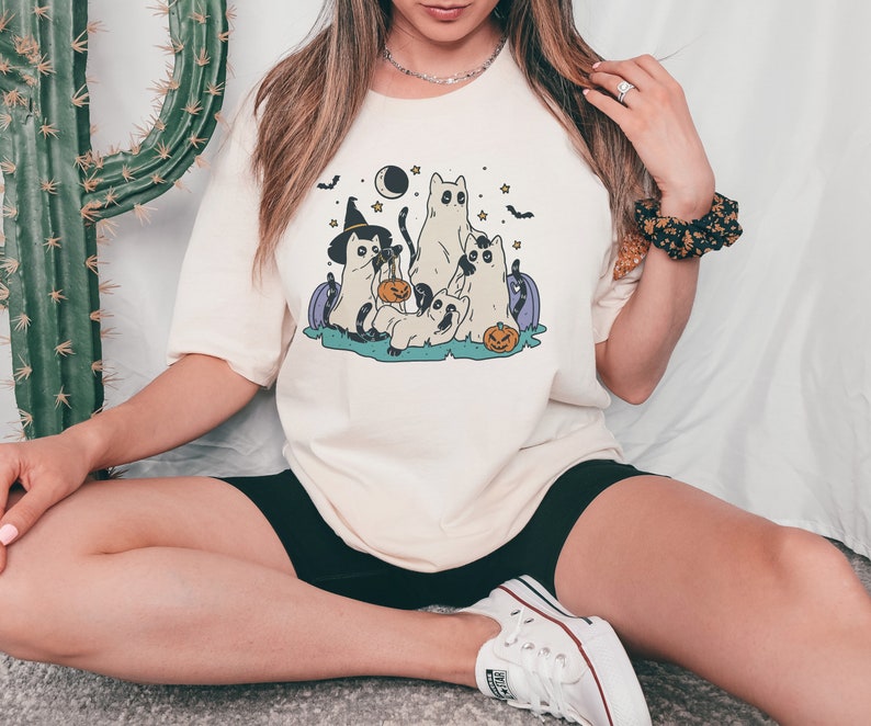 May include: A white t-shirt with a graphic of four cats dressed up for Halloween. The cats are wearing witch hats, holding pumpkins, and have stars and a crescent moon in the background.