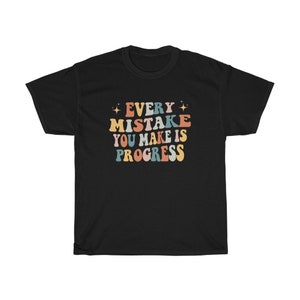 May include: Black t-shirt with the text "EVERY MISTAKE YOU MAKE IS PROGRESS" in colorful, retro-style lettering. The design includes stars and a vintage font, perfect for casual wear.